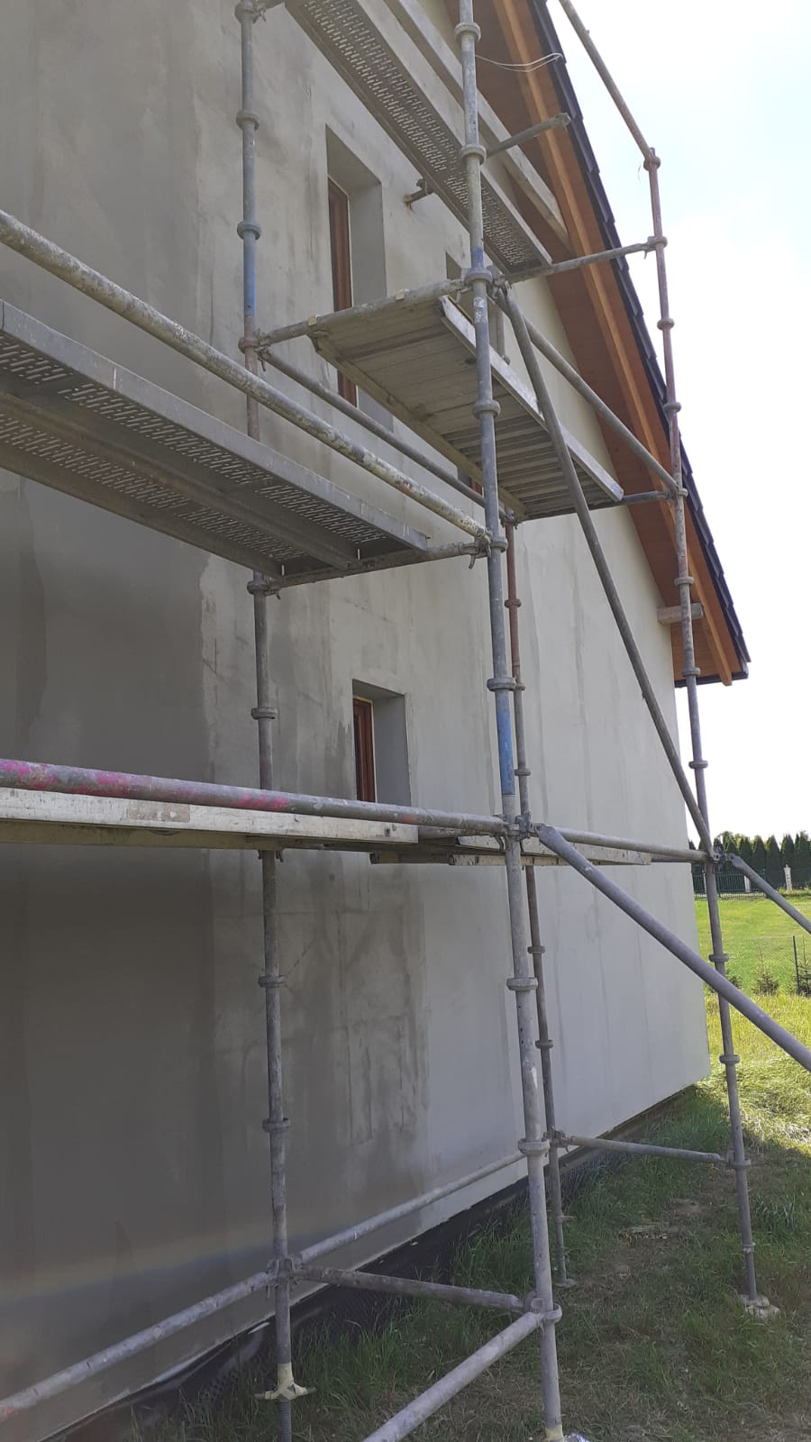 Ściana budynku w trakcie szpachlowania, widoczne rusztowanie i ślady po nałożonym materiale, okna z drewnianymi ramami.