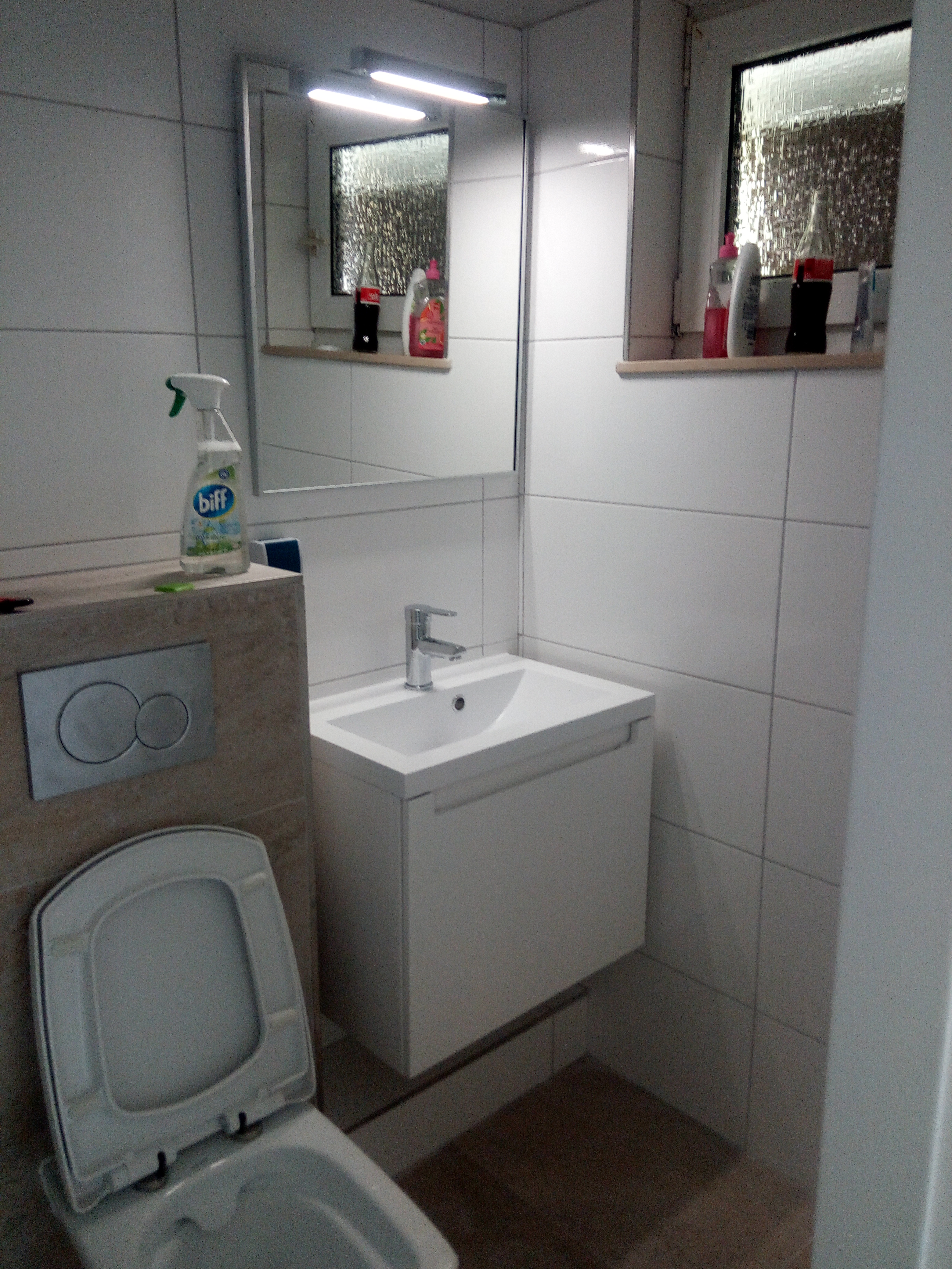 Biała umywalka z szafką i chromowaną baterią w wykafelkowanej łazience z widocznym WC i lustrem z oświetleniem. Na półce pod lustrem i na obudowie WC stoją środki czystości.