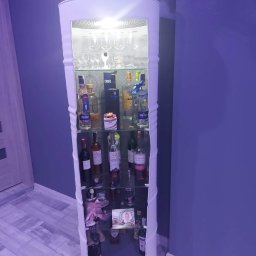 Errementari Adrian Leśniak - Barek w kształcie beczki z białym wykończeniem, podświetlony od wewnątrz, wypełniony butelkami alkoholu i szklankami, stojący w rogu pokoju z fioletowymi ścianami i jasną podłogą.