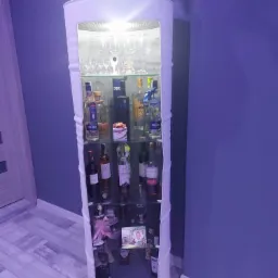 Barek w kształcie beczki z białym wykończeniem, podświetlony od wewnątrz, wypełniony butelkami alkoholu i szklankami, stojący w rogu pokoju z fioletowymi ścianami i jasną podłogą.