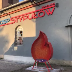 Budynek OSP z czerwonym napisem OSP Stypułów i rokiem założenia 1946, przed budynkiem czerwona metalowa rzeźba w kształcie płomienia, wypełniona kwiatami.