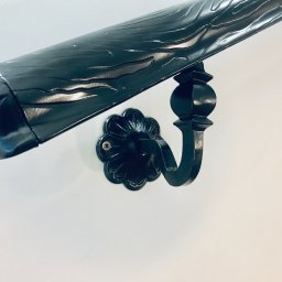 Errementari Adrian Leśniak - Czarna, metalowa poręcz schodowa z dekoracyjnym, kwiatowym mocowaniem przytwierdzona do białej ściany.