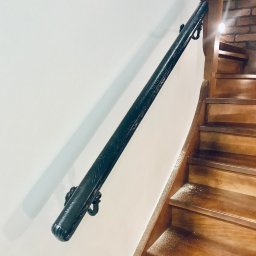 Errementari Adrian Leśniak - Czarna, zdobiona, metalowa poręcz zamocowana do białej ściany wzdłuż drewnianych schodów.