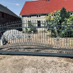 Przesuwna brama wjazdowa z kutymi elementami dekoracyjnymi, szara, z ząbkowanymi zakończeniami prętów, na tle domu z czerwoną dachówką i kwitnących słoneczników.