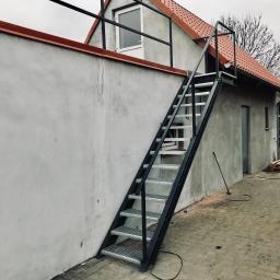 Errementari Adrian Leśniak - Zewnętrzne metalowe schody z kratowymi stopniami, prowadzące na dach budynku z czerwoną dachówką, widoczne na tle szarej ściany i kostki brukowej.