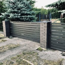 Errementari Adrian Leśniak - Nowoczesna brama wjazdowa z poziomymi panelami w kolorze grafitowym, osadzona na ceglanych słupkach z ciemnymi nakrywami, na tle zielonych drzew i trampoliny.