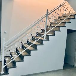 Errementari Adrian Leśniak - Schody wewnętrzne z metalową, białą balustradą o dekoracyjnym wzorze liści i kwiatów, widok z dołu, w tle jasne ściany i drzwi.