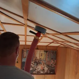 Aplikacja pianki PUR na poddaszu z drewnianą konstrukcją, widoczna czerwona rura aplikatora i obraz na ścianie w tle.