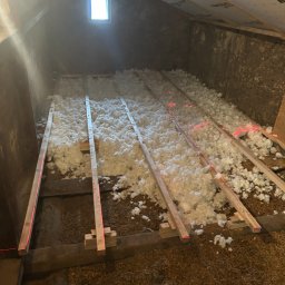 HANSLIK JAROSŁAW JARTRANS - Poddasze w trakcie izolacji: biała wełna mineralna między drewnianymi legarami, wyznaczona poziomica laserowa, małe okno w szczycie.