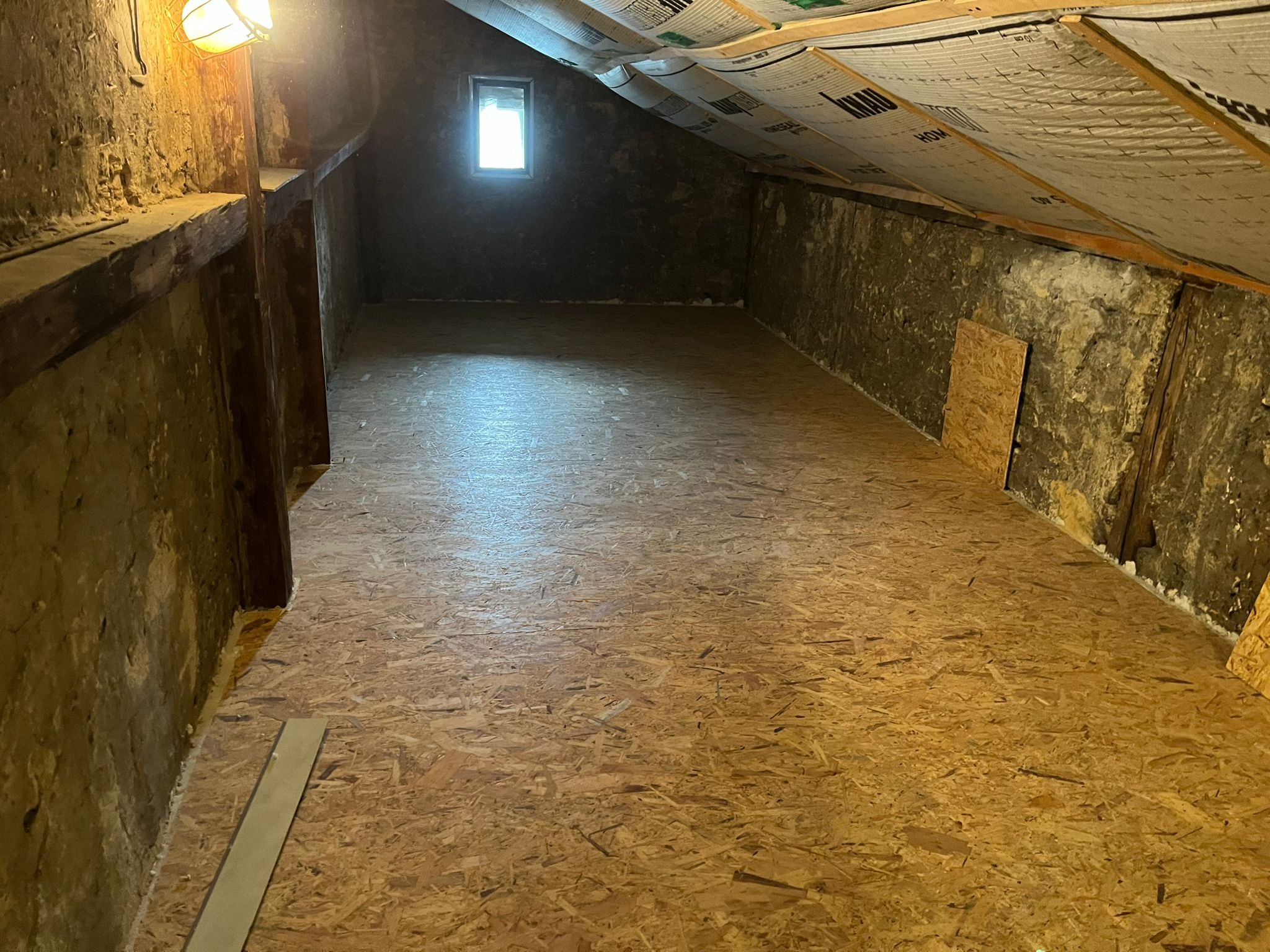 Poddasze po ociepleniu, widoczne płyty OSB na podłodze, ocieplenie między krokwiami dachu z widoczną folią paroizolacyjną, fragment ściany z resztkami starego tynku i małe okno w szczycie.