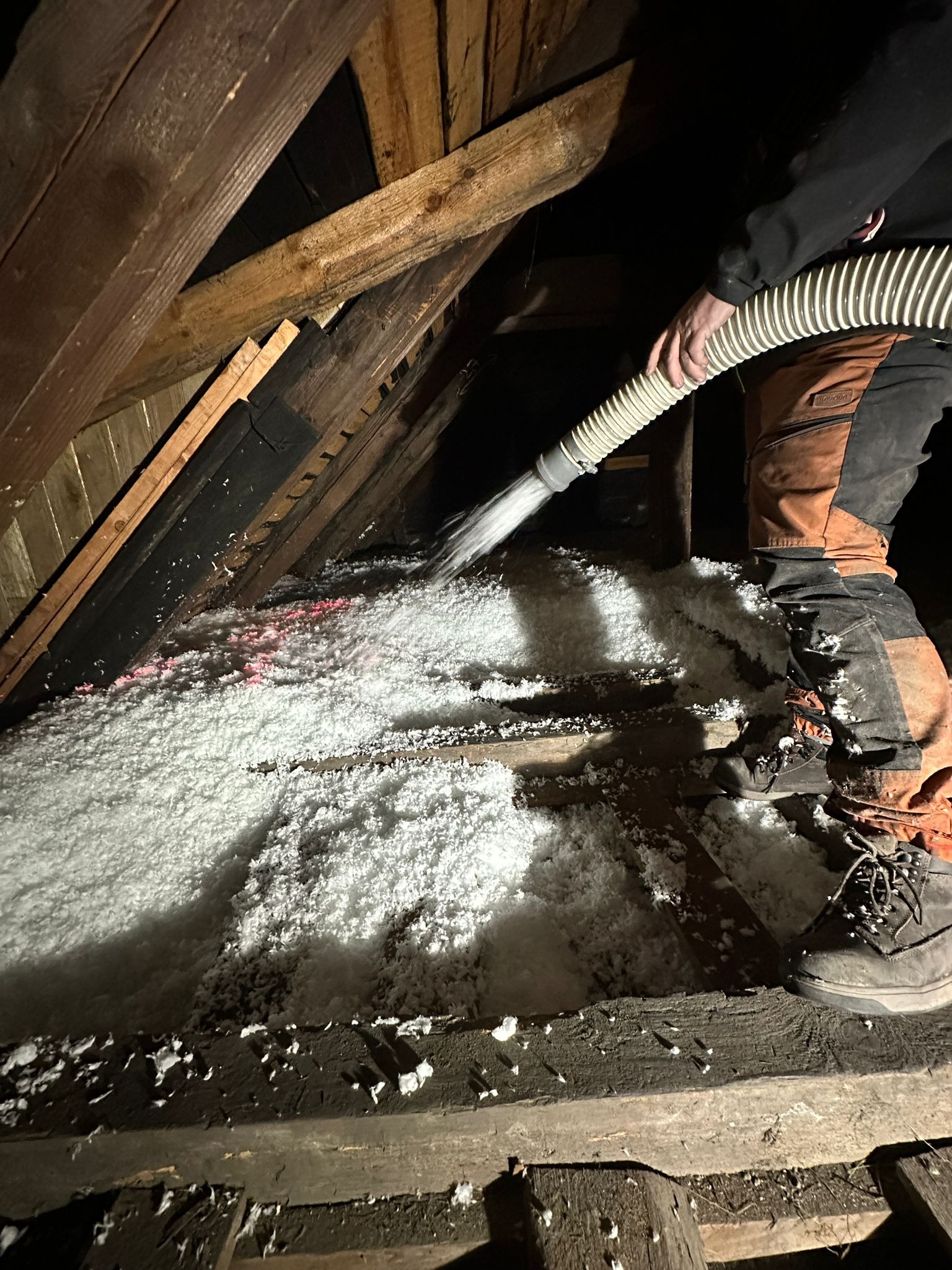 Aplikacja białej izolacji celulozowej w przestrzeni poddasza z użyciem węża, widoczne drewniane elementy konstrukcji dachu i fragment roboczego stroju osoby wykonującej pracę.