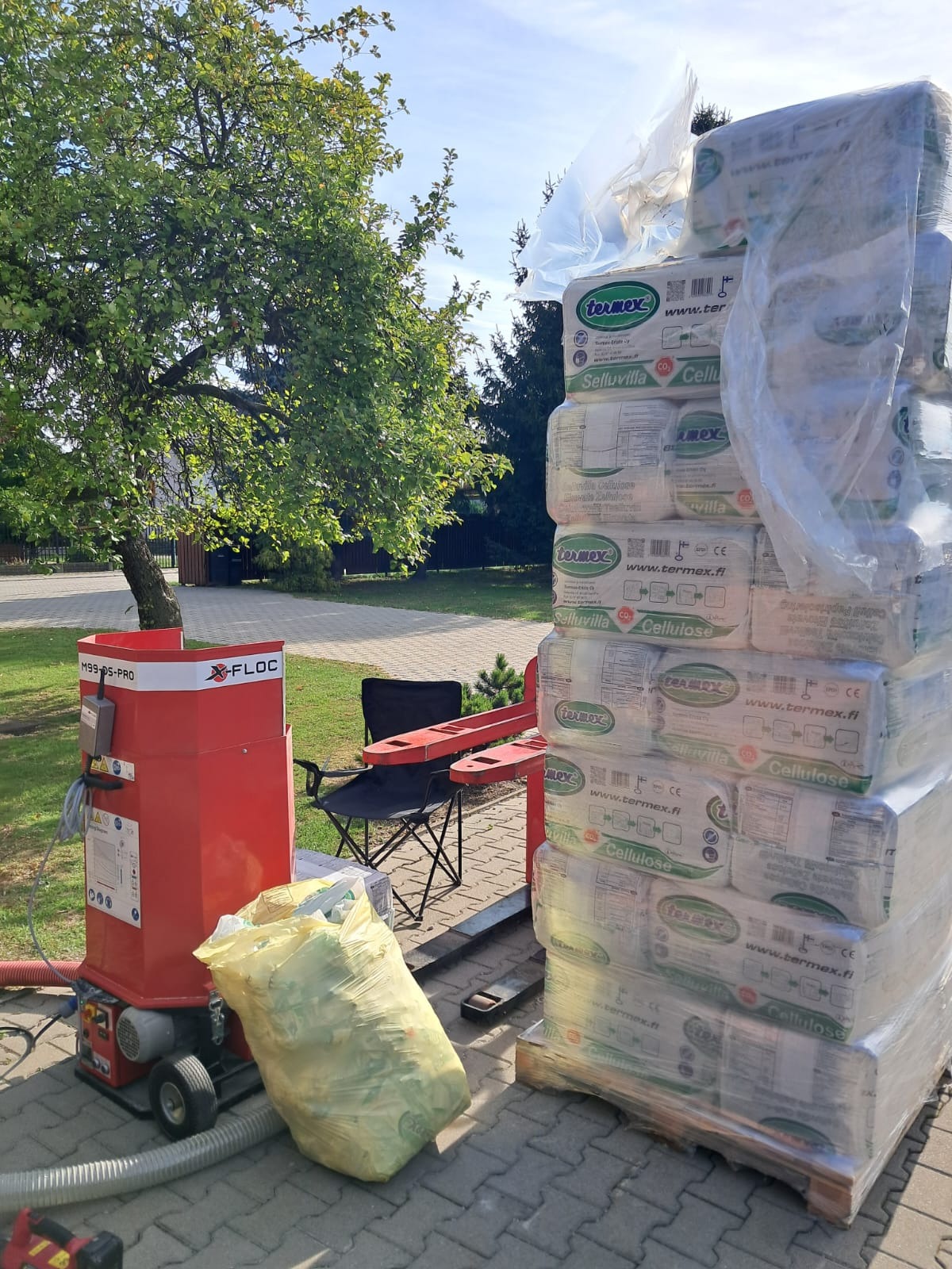 Stos sprasowanej celulozy marki Termex Selluvilla na palecie w Chudowie, obok czerwona maszyna X-Floc M99-DS-PRO i worek z odpadami, na tle trawnik i drzewa.