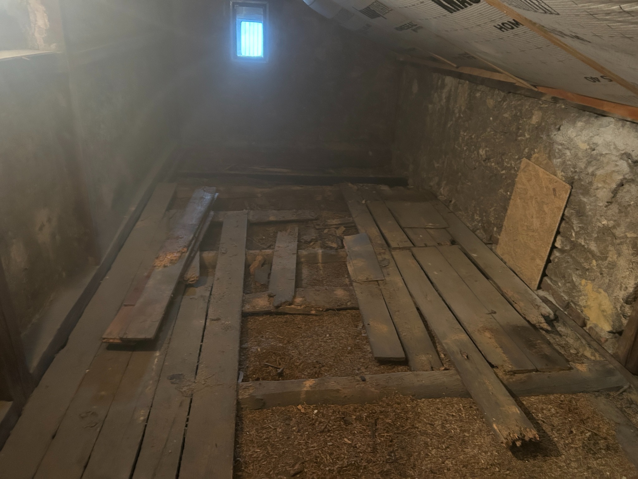 Poddasze z odsłoniętą konstrukcją drewnianą podłogi, widoczne fragmenty izolacji na dachu i ściany z kamienia, małe okno dachowe w tle.