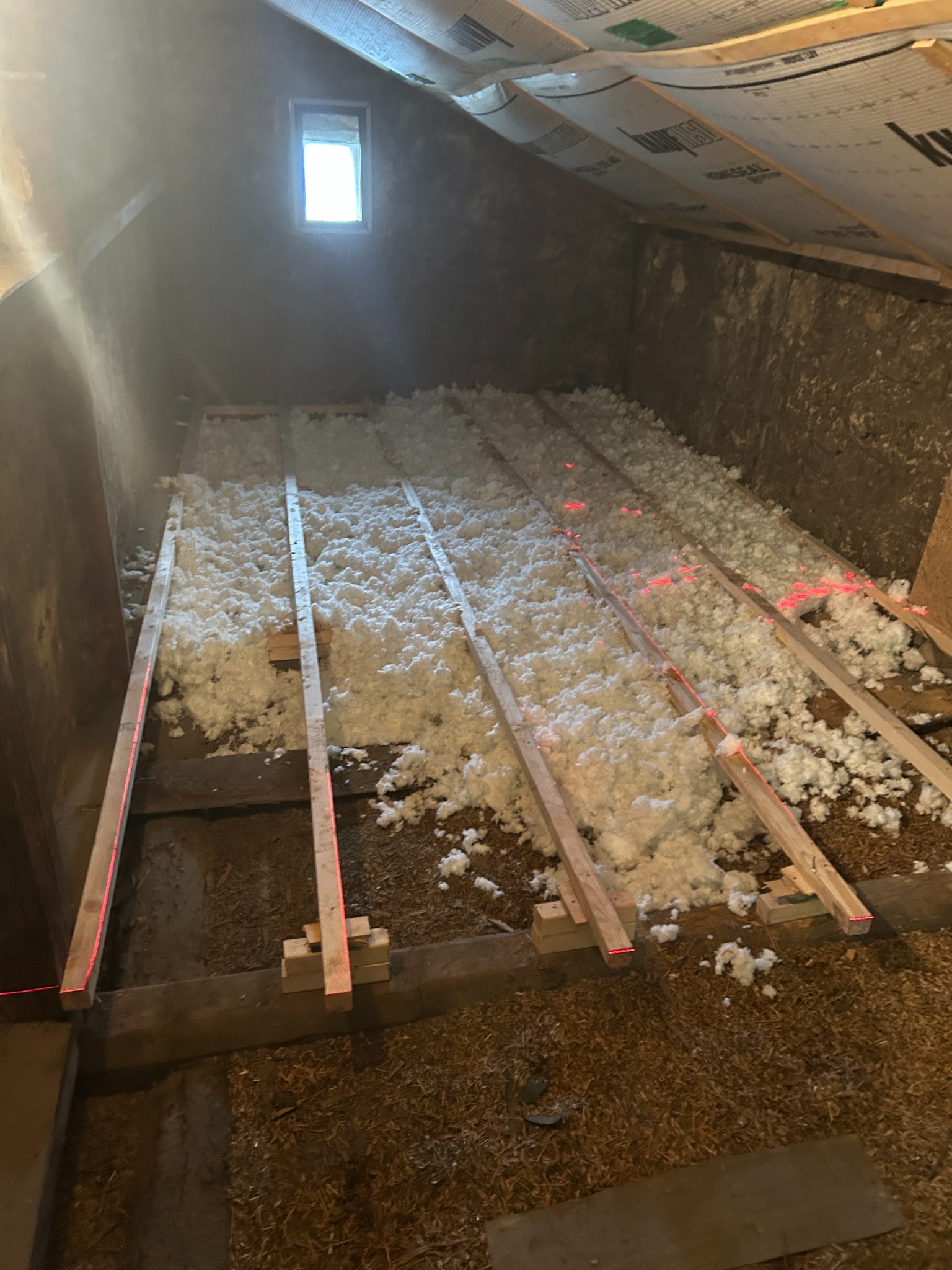 Poddasze w trakcie izolacji: biała wełna mineralna między drewnianymi legarami, wyznaczona poziomica laserowa, małe okno w szczycie.
