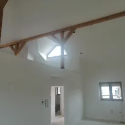 Wnętrze pomieszczenia po remoncie z drewnianą konstrukcją stropu i oknem dachowym, świeżo pomalowane ściany na biało, widoczne ślady po instalacjach elektrycznych.