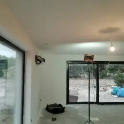 Wnętrze w trakcie remontu z dużymi oknami przesuwnymi, widoczny surowy stan ścian, lampa wisząca na kablu, materiały budowlane i drabina teleskopowa.