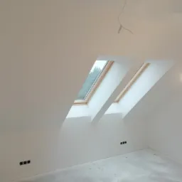 Poddasze po remoncie z dwoma oknami dachowymi, świeżo pomalowane na biało, widoczne gniazdka elektryczne i surowy beton na podłodze, wisząca żarówka.