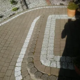 Ułożona z kostki brukowej ścieżka ogrodowa z białymi elementami dekoracyjnymi i obrzeżem z szarych, okrągłych kamieni; w tle widoczny fragment ogrodu z roślinami i jasnymi kamieniami.