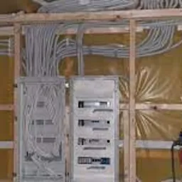 Rozdzielnice elektryczne z licznymi przewodami w szarych peszlach, zamontowane na drewnianej konstrukcji w surowym stanie.