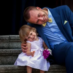 Uśmiechnięty mężczyzna w niebieskim garniturze i żółtej muszce obejmuje siedzącą na schodach dziewczynkę w jasnej sukience trzymającą bukiecik fioletowych kwiatów.