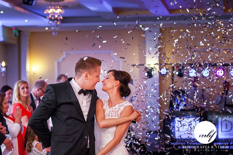 Para młoda, on w czarnym smokingu z muchą, ona w białej sukni ślubnej z koronkową górą, obejmująca się na parkiecie w strugach konfetti, w tle oświetlenie sceniczne i goście.