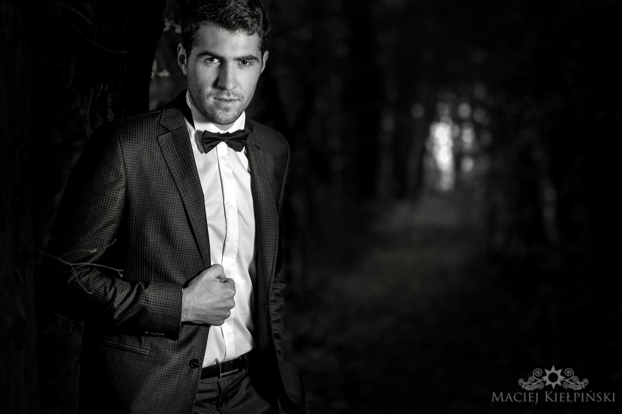 Czarno-białe portretowe ujęcie mężczyzny w garniturze i muszce na tle rozmytego, leśnego krajobrazu.