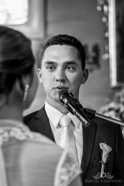 Czarno-białe zbliżenie mężczyzny w garniturze, z łzą spływającą po policzku, mówiącego do mikrofonu podczas ceremonii ślubnej; widoczna panna młoda w tle.