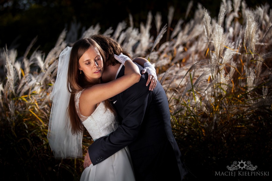 Panna młoda w koronkowej sukni i welonie, z zamkniętymi oczami, obejmuje pana młodego w ciemnym garniturze na tle wysokich traw w złotym świetle.