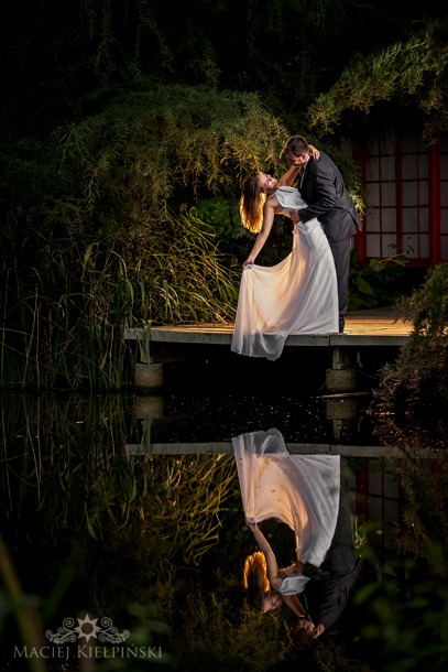 Para młoda w tańcu na drewnianym pomoście nad wodą, z odbiciem w tafli i zielenią w tle, uchwycona w romantycznym oświetleniu podczas sesji poślubnej.