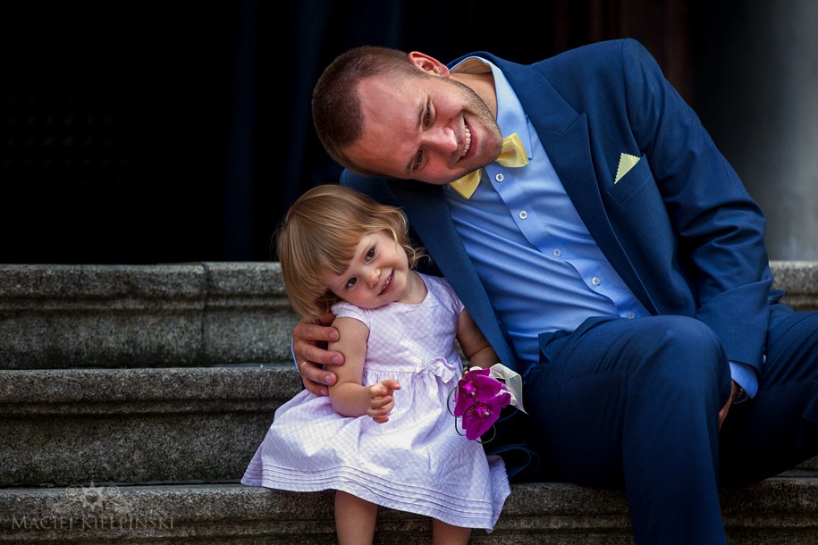 Uśmiechnięty mężczyzna w niebieskim garniturze i żółtej muszce obejmuje siedzącą na schodach dziewczynkę w jasnej sukience trzymającą bukiecik fioletowych kwiatów.
