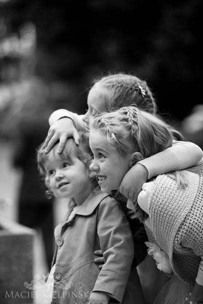Czarno-białe zdjęcie trzech dziewczynek, obejmujących się i spoglądających w dal z uśmiechem, włosy zaplecione w warkocze, delikatne oświetlenie.