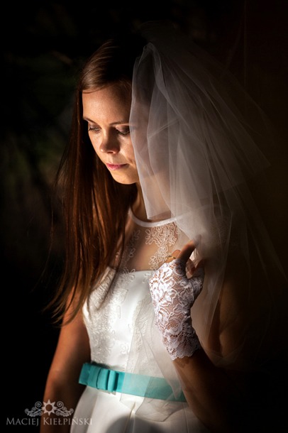 Portret młodej kobiety w białej sukni z turkusową wstążką, w welonie i koronkowych rękawiczkach, spoglądającej w dół w miękkim, bocznym oświetleniu.