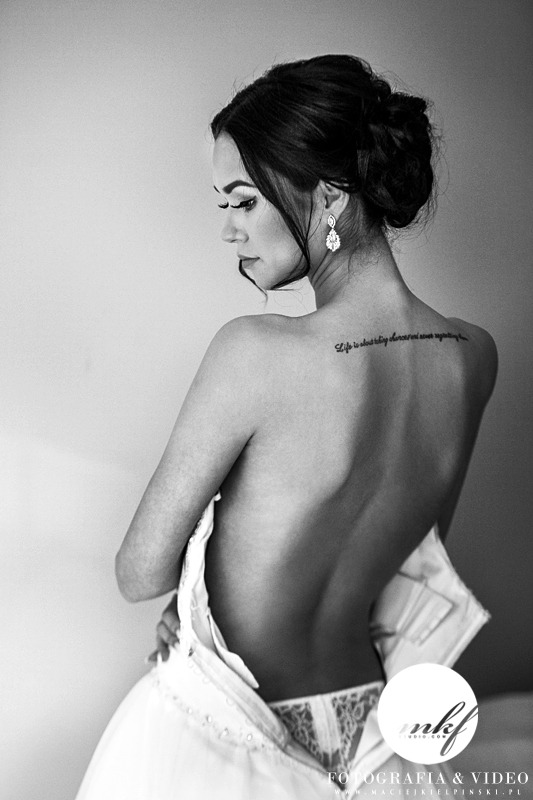 Czarno-białe zdjęcie panny młodej z odsłoniętymi plecami, częściowo ubrana w białą suknię ślubną, z widocznym tatuażem na plecach i wiszącymi kolczykami.