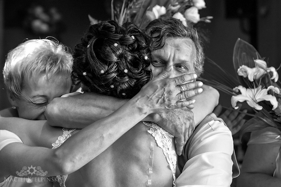 Czarno-białe zdjęcie wzruszonego ojca, zakrywającego usta dłonią z widocznymi żyłami, obejmującego pannę młodą w sukni z koronkowymi ramiączkami oraz kobietę, prawdopodobnie matkę, podczas...