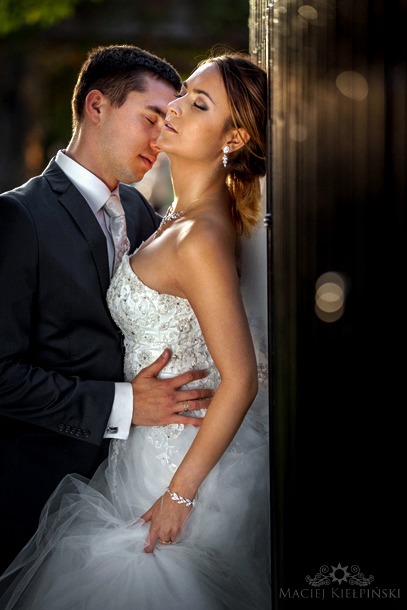 Para młoda w objęciach, panna młoda opiera się o ciemną konstrukcję, zamknięte oczy, delikatne oświetlenie podkreśla detale sukni i biżuterii.