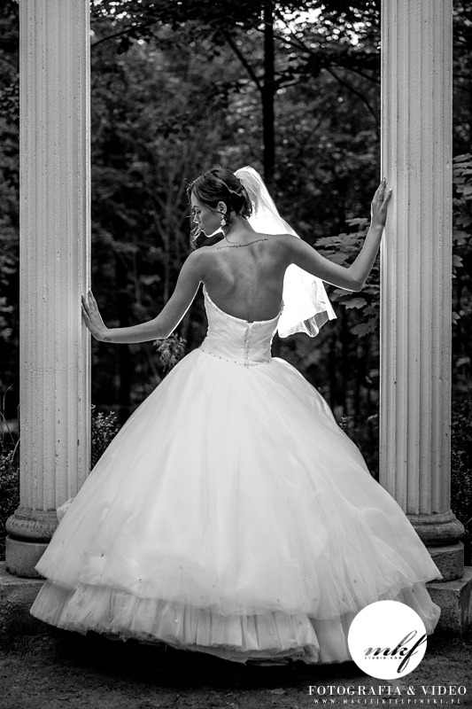 Czarno-białe zdjęcie panny młodej w sukni balowej, stojącej tyłem, opierającej się o kolumny w plenerze, z widocznym tatuażem na plecach i delikatnym welonem.