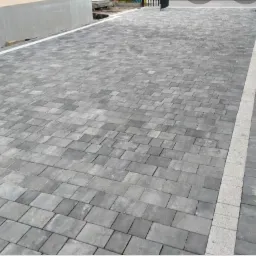 Ułożona szara kostka brukowa w regularny wzór, z jasną linią oddzielającą od chodnika, widoczna zagęszczarka w tle.