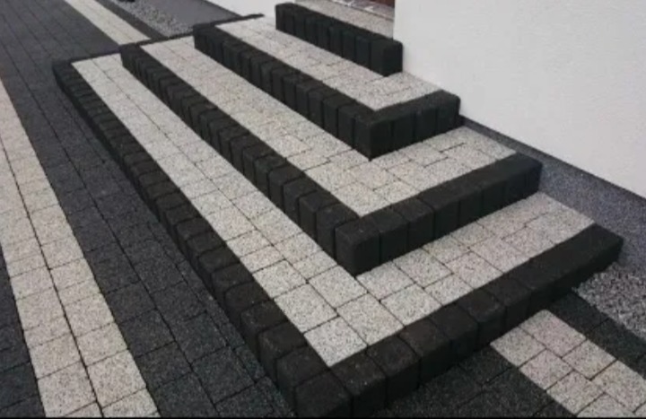 Schody zewnętrzne wyłożone szarą kostką granitową z czarnym obramowaniem, prowadzące do białej ściany budynku.