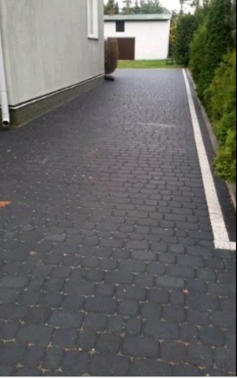 Ciemnoszara kostka brukowa ułożona wzdłuż budynku z białym paskiem oddzielającym chodnik od trawnika, w tle budynek gospodarczy.