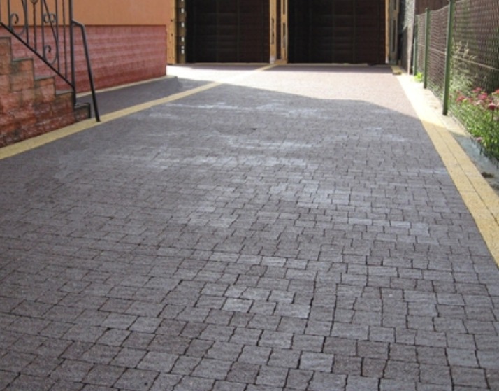 Ciemnoszara kostka granitowa ułożona na podjeździe, z żółtym obrzeżem, prowadząca do garażu z metalowymi drzwiami, widok z perspektywy