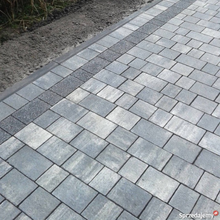 Ułożona szara kostka brukowa w prostokątnym wzorze z obramowaniem z drobnego grysu, widoczne świeżo usypane ziemne obrzeże.