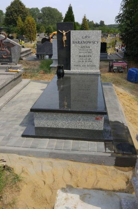 Nowy, granitowy nagrobek z czarnym wazonem na błyszczącej płycie, otoczony jasnoszarą kostką brukową na cmentarzu, z widocznym generatorem w tle.