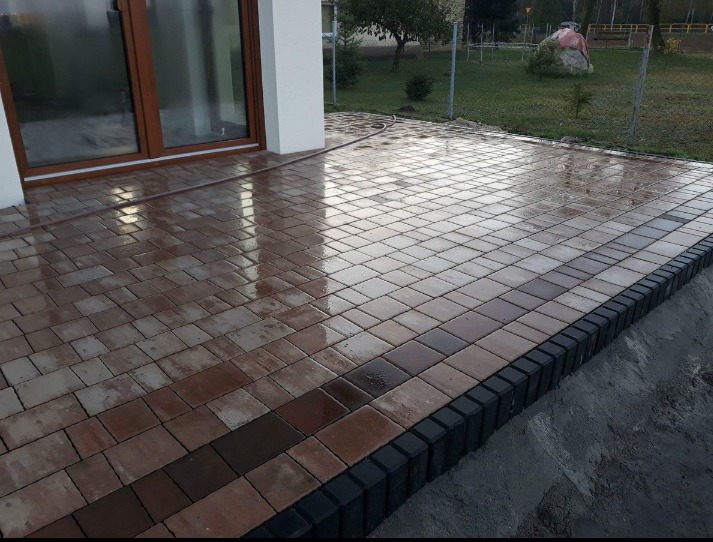 Świeżo ułożona, mokra kostka brukowa w odcieniach brązu i szarości, obramowana czarnym obrzeżem, przylegająca do jasnego budynku z brązowymi drzwiami tarasowymi, widoczny fragment trawnika w tle.