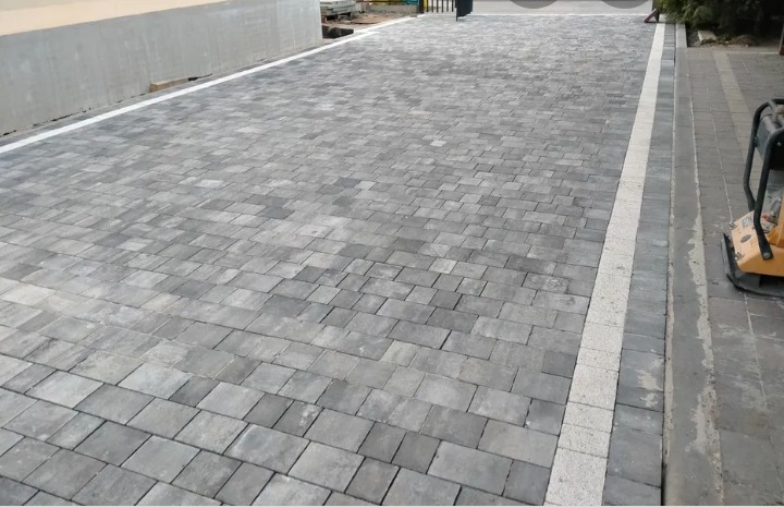 Ułożona szara kostka brukowa w regularny wzór, z jasną linią oddzielającą od chodnika, widoczna zagęszczarka w tle.