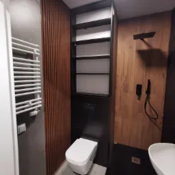 Nowoczesna łazienka z drewnianymi akcentami: pionowe panele na ścianie, kabina prysznicowa walk-in z czarną armaturą, podwieszana toaleta i grzejnik drabinkowy.