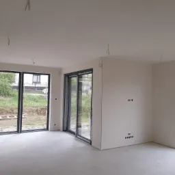 Wnętrze w stanie deweloperskim, ściany przygotowane pod malowanie, widoczne wyprowadzenia elektryczne i przewody oświetleniowe zwisające z sufitu, duże okna przesuwne z widokiem na zewnątrz.