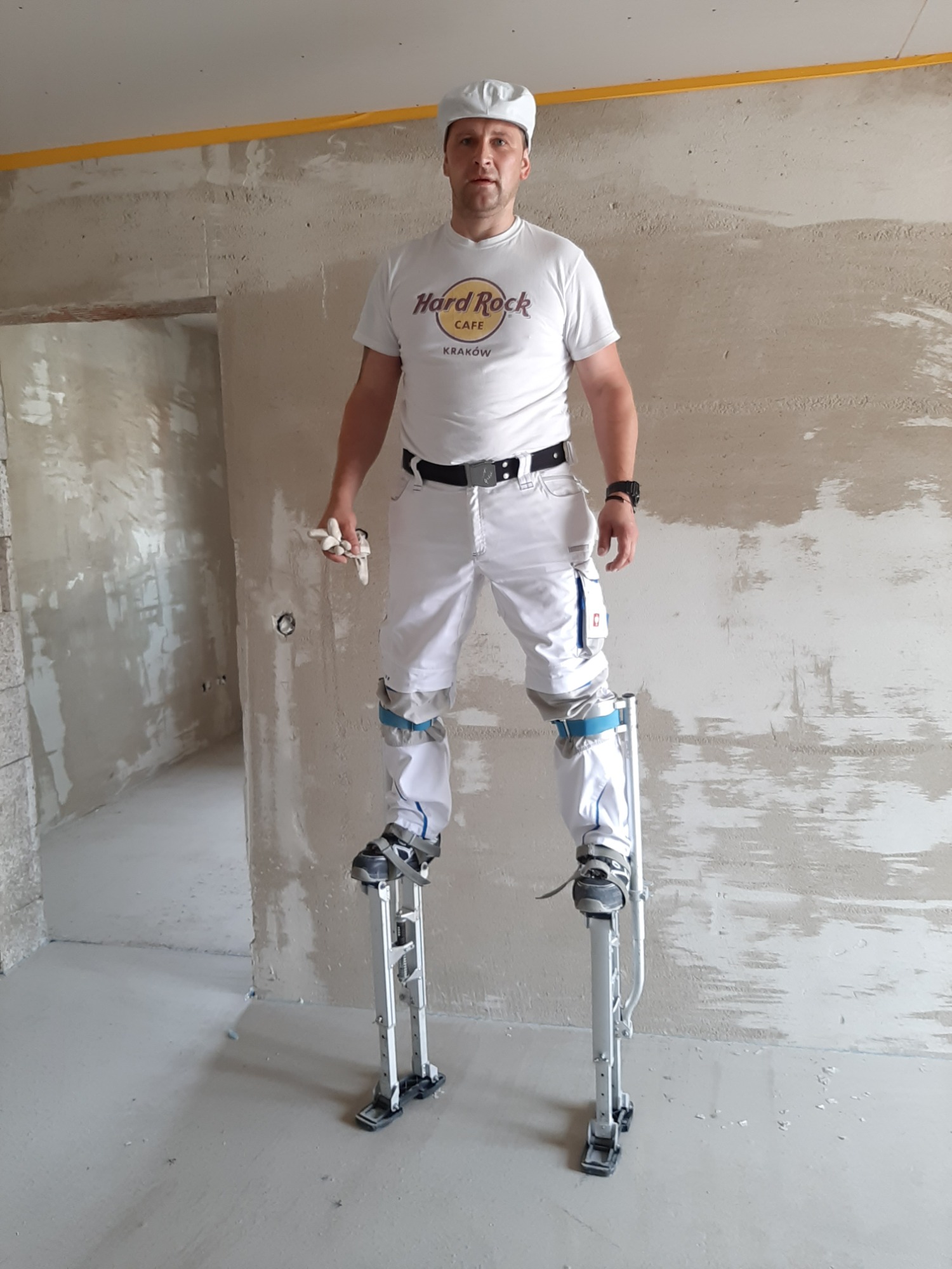 Pracownik w białym stroju roboczym i na szczudłach, przygotowuje ściany do dalszych prac wykończeniowych w surowym pomieszczeniu z widocznymi śladami szpachlowania.