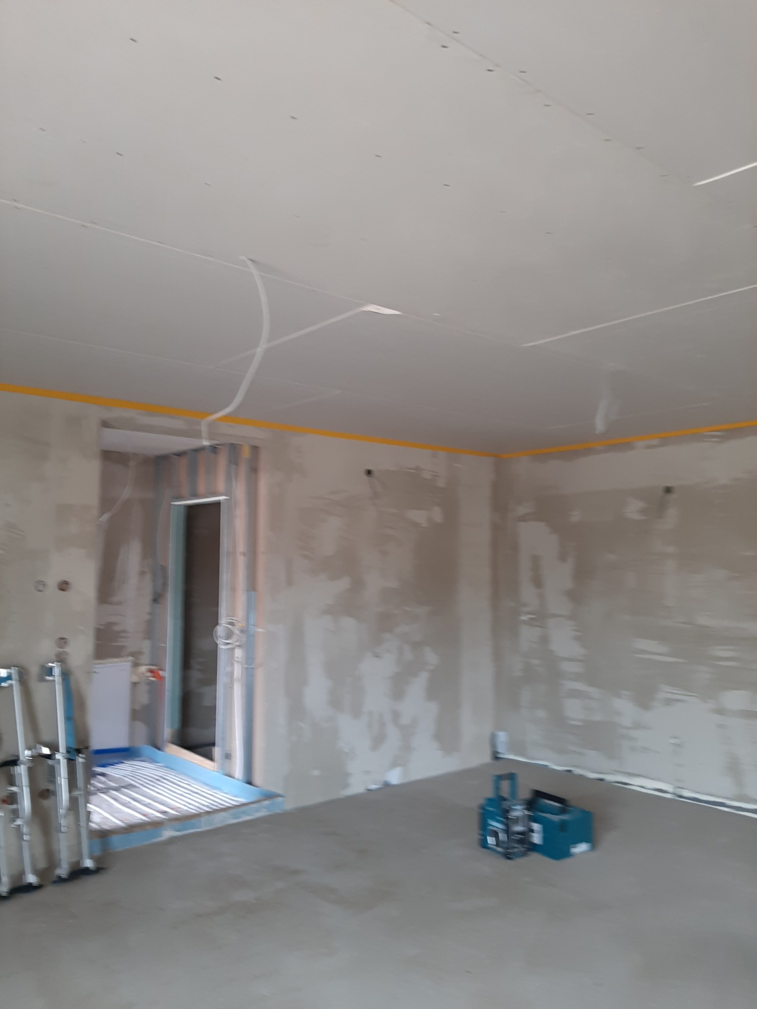 Wnętrze w trakcie remontu, widoczne ściany po szpachlowaniu, sufit z płyt kartonowo-gipsowych, taśma malarska wzdłuż krawędzi, narzędzia do szpachlowania i drabiny na podłodze.