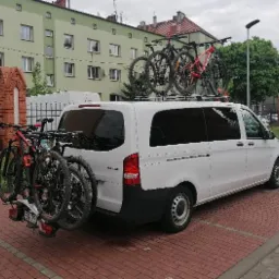 Biały bus z bagażnikiem rowerowym na haku i trzema rowerami na dachu, zaparkowany na chodniku z czerwonej kostki, z budynkiem mieszkalnym w tle.