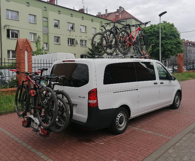 Biały bus z bagażnikiem rowerowym na haku i trzema rowerami na dachu, zaparkowany na chodniku z czerwonej kostki, z budynkiem mieszkalnym w tle.